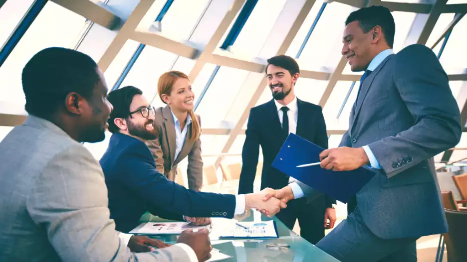 A group of diverse professionals in a modern office space, shaking hands and collaborating over documents, with large windows letting in ample sunlight.